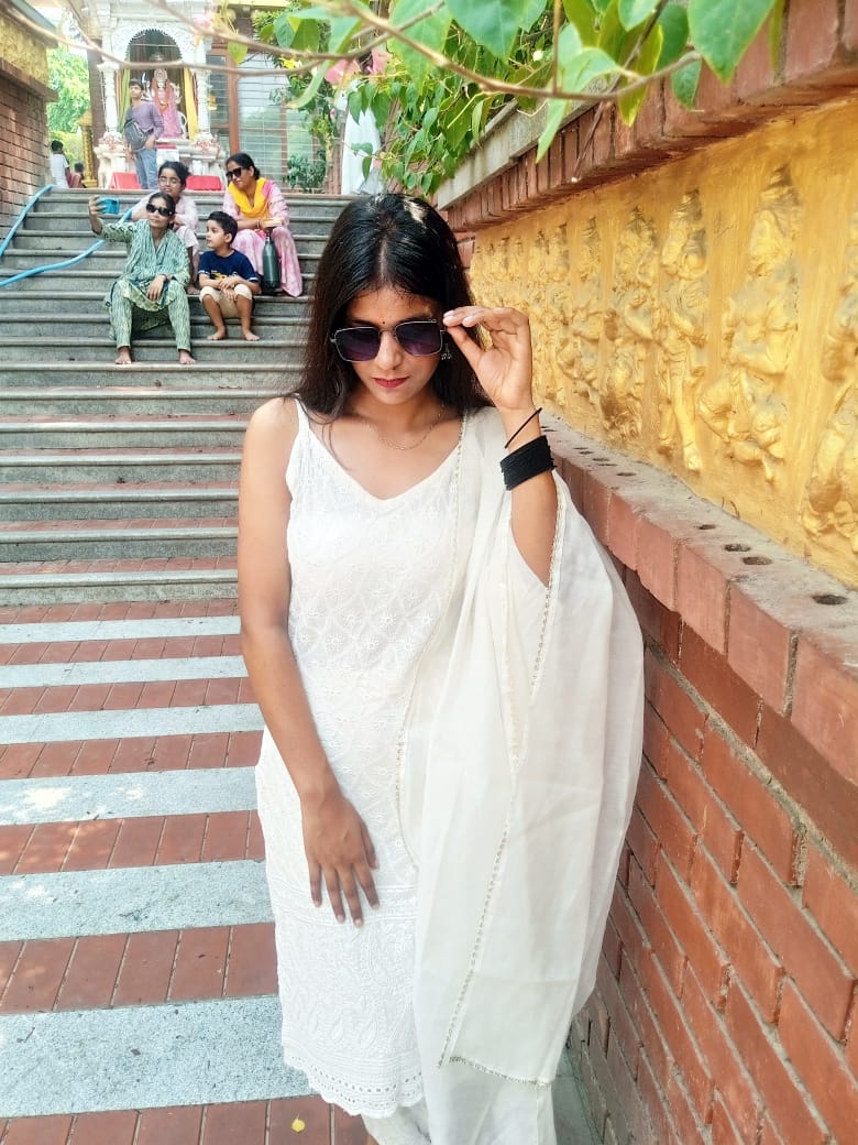 Saumya looking down and adjusting sunglasses in a white suit by the golden temple wall