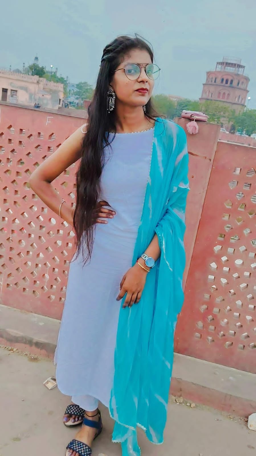 Saumya in a lavender suit with a turquoise dupatta and round glasses, in front of a red-stone lattice wall